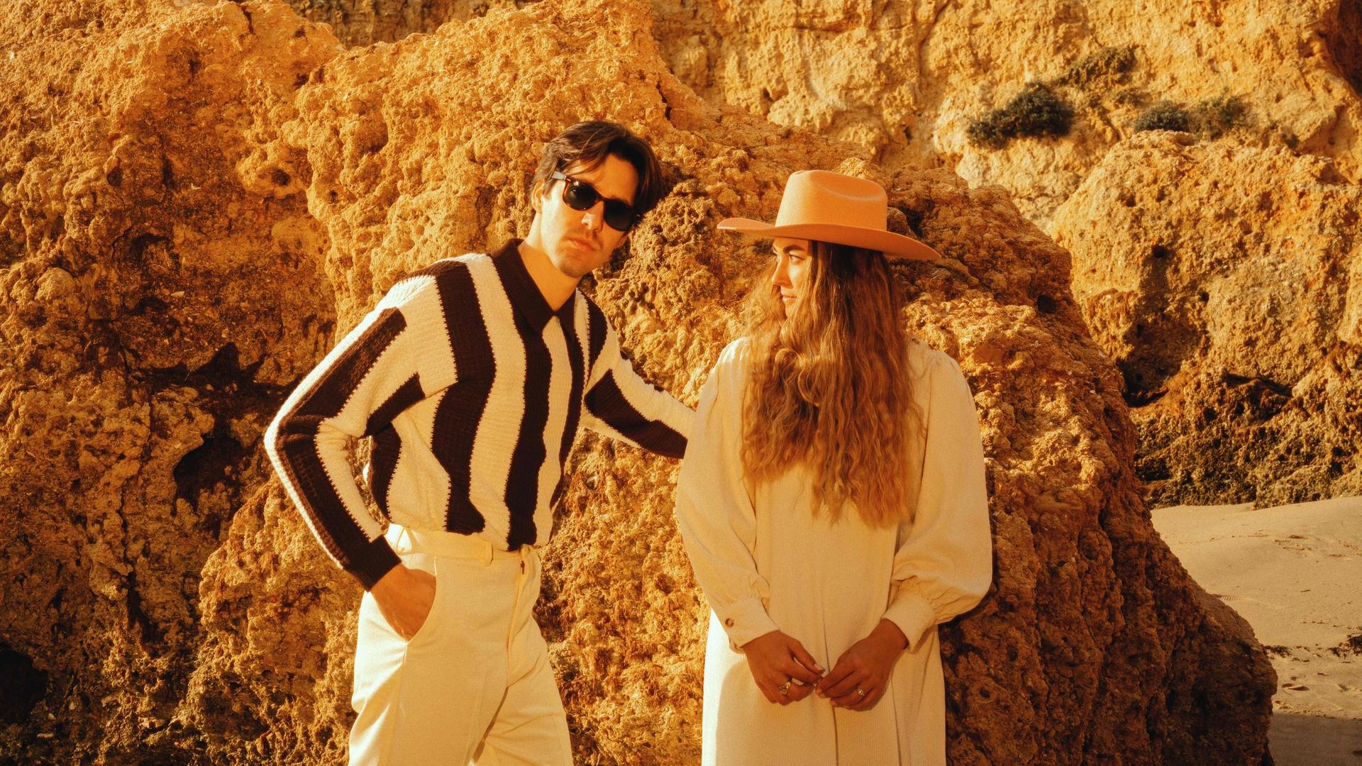 Ein Mann und eine Frau mit langen Haaren und Hut stehen vor einem sandigen Felsen eng beieinander.