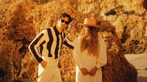 Ein Mann und eine Frau mit langen Haaren und Hut stehen vor einem sandigen Felsen eng beieinander.