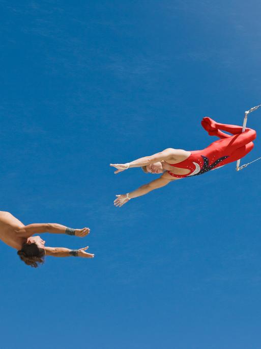 Zwei Trapezkünstler vor blauem Himmel. Sie strecken sich die Arme gegenseitig zu.
