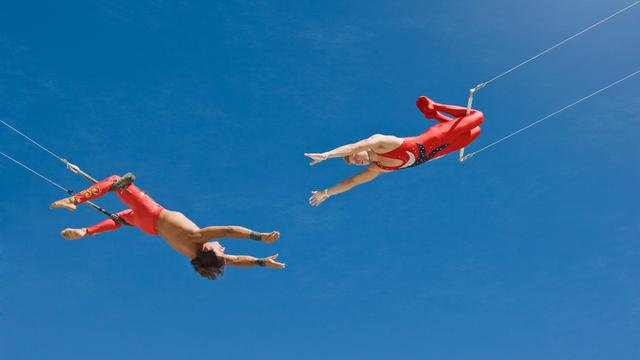 Zwei Trapezkünstler vor blauem Himmel. Sie strecken sich die Arme gegenseitig zu.
