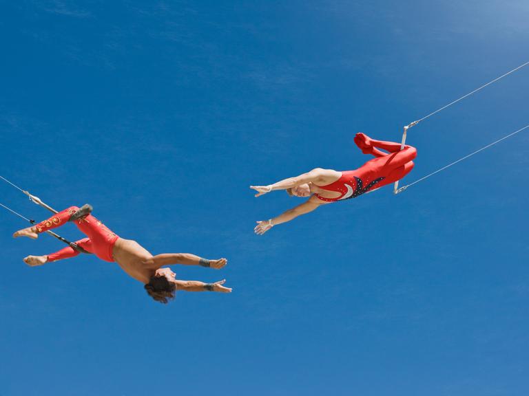 Zwei Trapezkünstler vor blauem Himmel. Sie strecken sich die Arme gegenseitig zu.