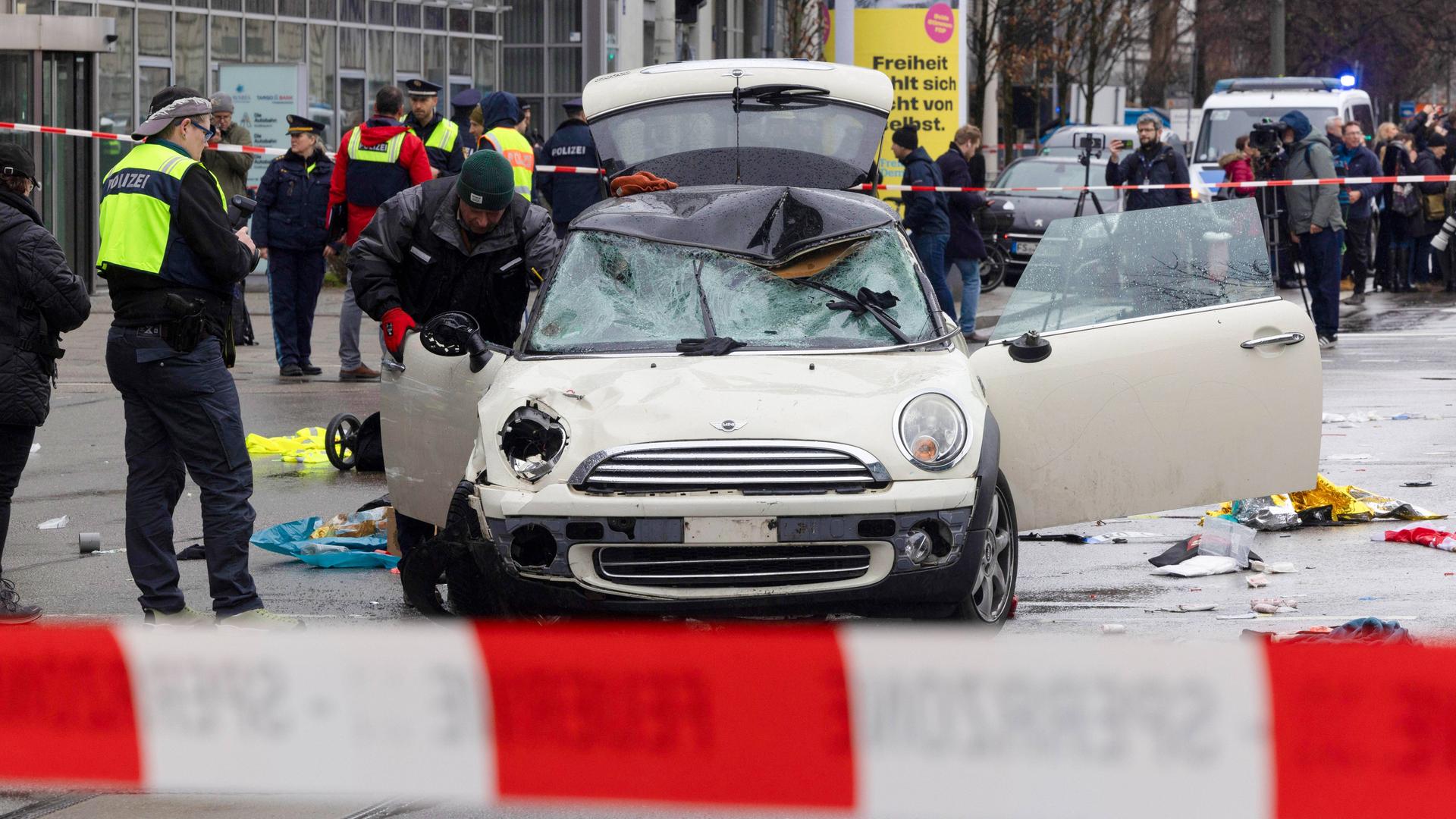 Polizisten stehen um den verbeulten weißen Minicooper herum, den man von vorne sieht. Im Vordergrund ein rot-weiß gestreiftes Flatterband. 