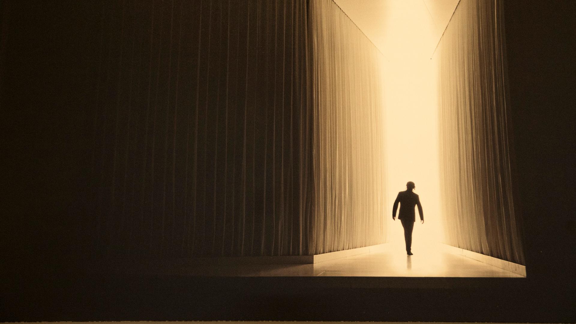 Bühnenbild: dunkle Wände links und rechts führen einen Weg zum Licht auf den sich die Silhouette einer Person abbildet