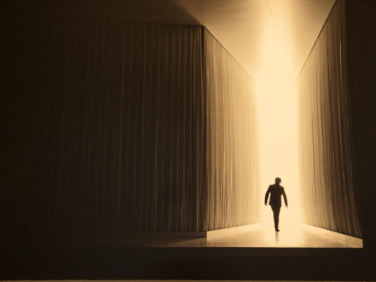 Bühnenbild: dunkle Wände links und rechts führen einen Weg zum Licht auf den sich die Silhouette einer Person abbildet