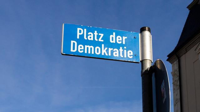Ein Straßenschild "Platz der Demokratie" steht in  Weimar. Im Hintergrund leuchtet der blaue Himmel. 