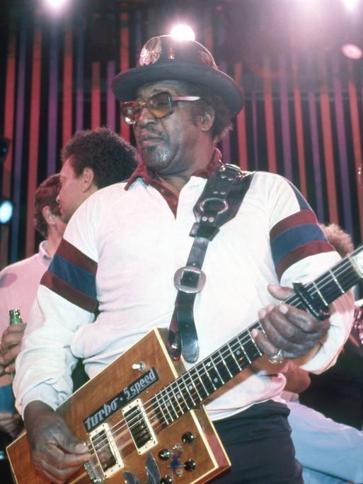 Der Musiker Bo Diddley trägt Hut und Brille und spielt eine kastenförmige Gitarre.