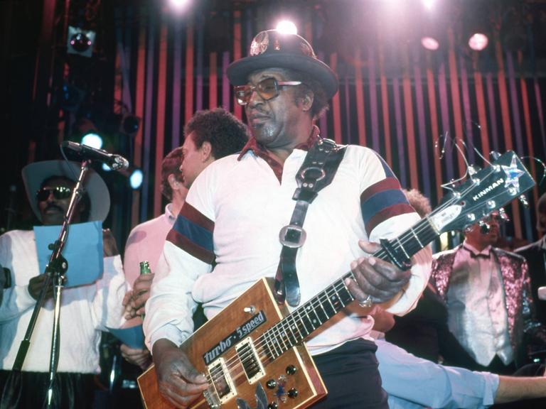 Der Musiker Bo Diddley trägt Hut und Brille und spielt eine kastenförmige Gitarre.