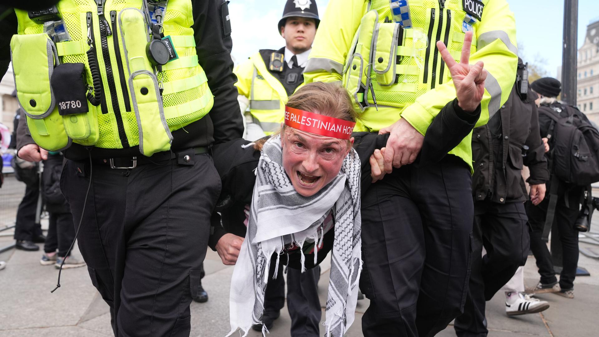 Polizeibeamte tragen eine Demonstrantin von einer Kundgebung gegen das Verbot von "Palestine Action" auf dem Trafalgar Square weg. Die Frau macht das Victory-Zeichen. Sie trägt ein Palästinensertuch.