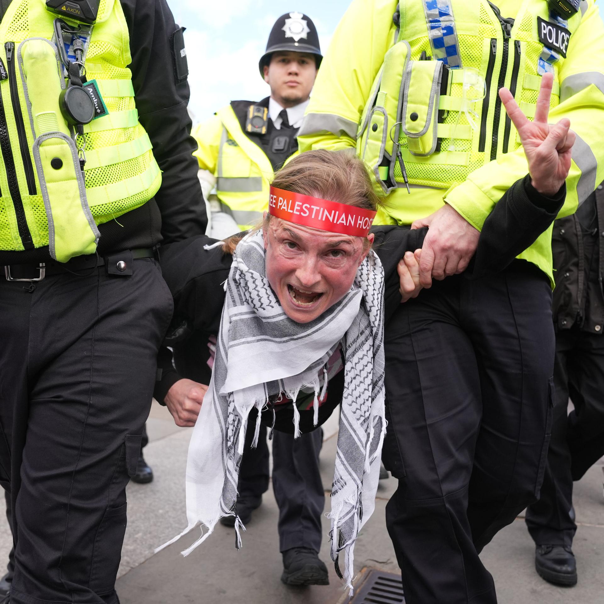 Polizeibeamte tragen eine Demonstrantin von einer Kundgebung gegen das Verbot von "Palestine Action" auf dem Trafalgar Square weg. Die Frau macht das Victory-Zeichen. Sie trägt ein Palästinensertuch.