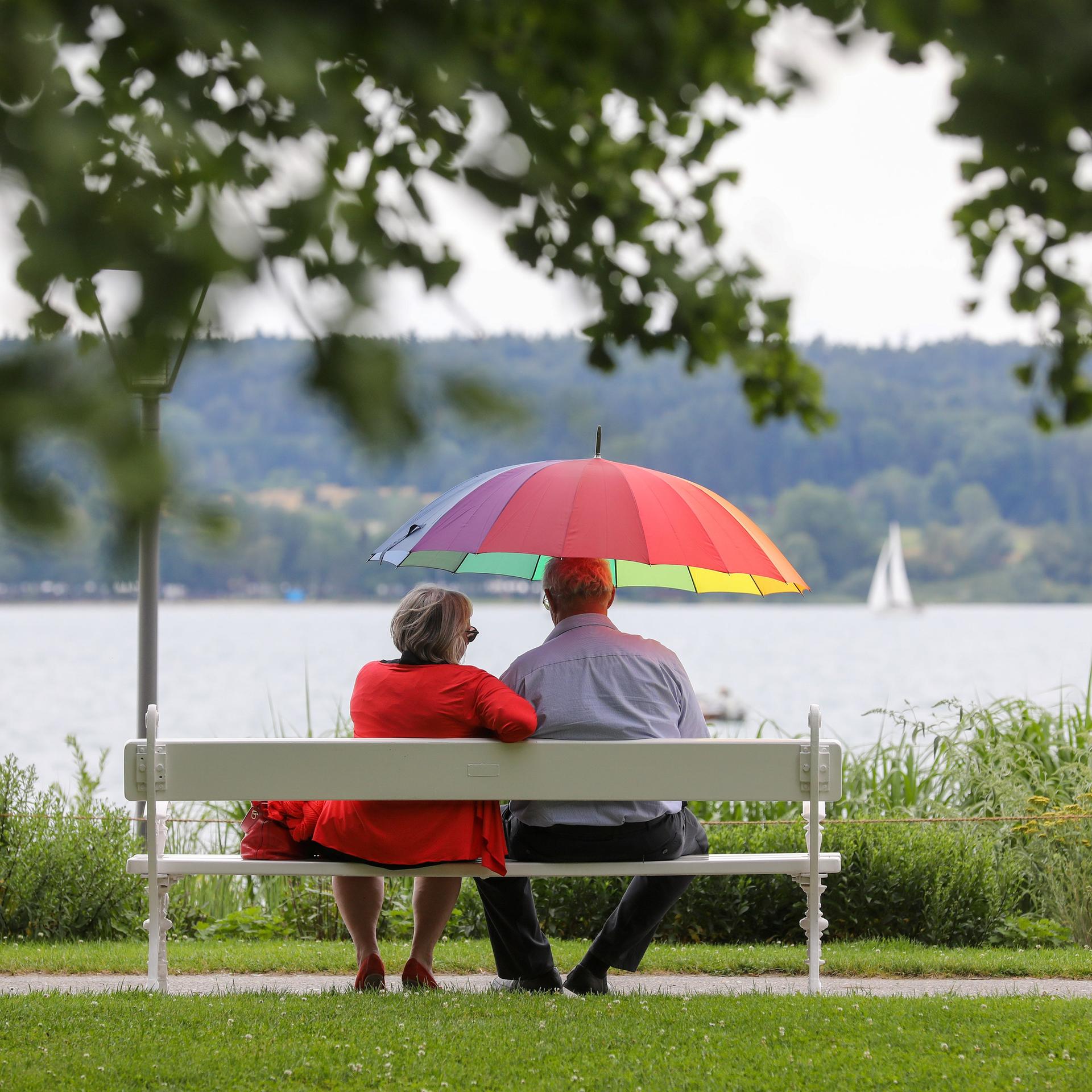 Ein Mann und eine Frau sitzen am Ufer des Bodensees mit einem Sonnenschirm in der Hand auf einer Bank.