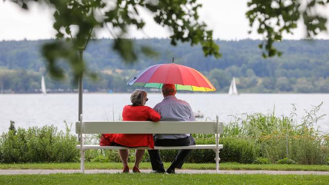 Ein Mann und eine Frau sitzen am Ufer des Bodensees mit einem Sonnenschirm in der Hand auf einer Bank.
