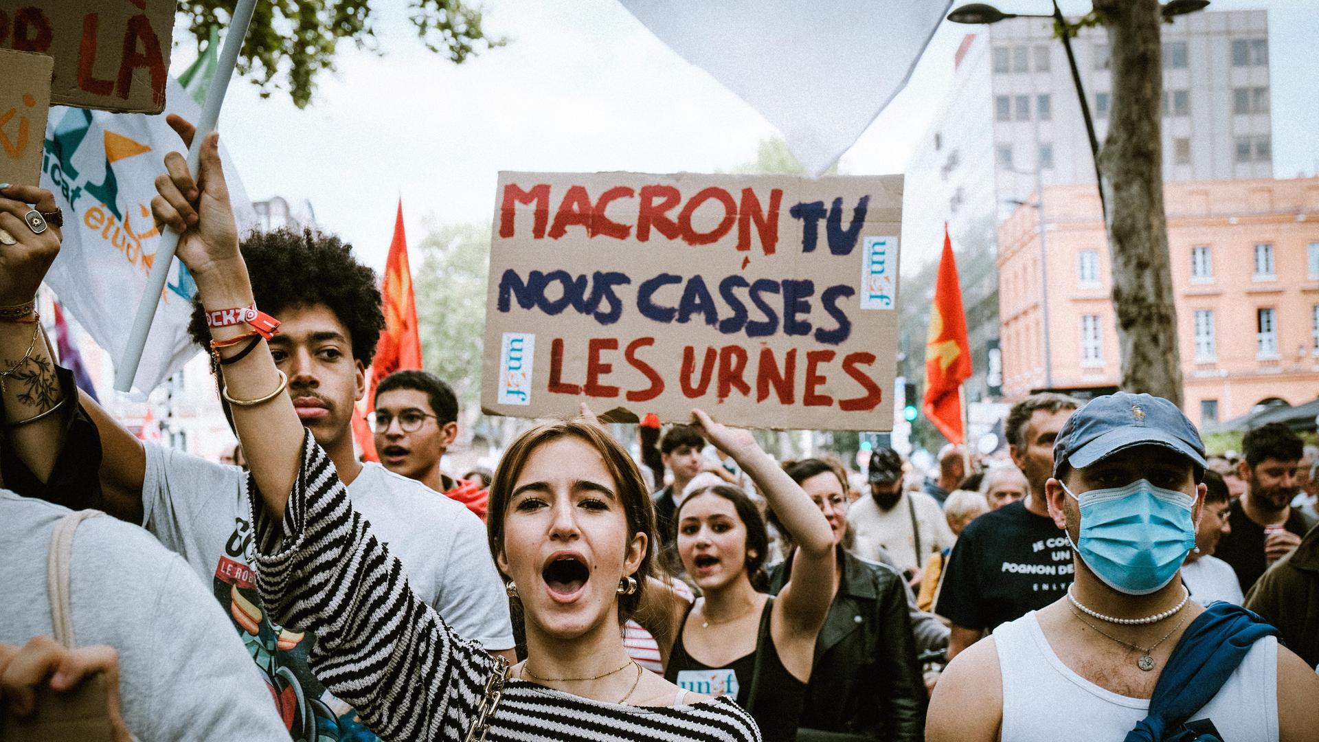 Demonstranten skandieren und halten ein Schild mit der Aufschrift „Macron, Tu Nous Casses Les Urnes“ ("Macron, du machst unsere Wahlurnen kaputt") in die Höhe.