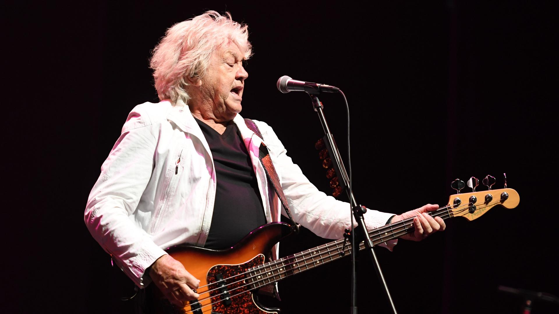 Der Musiker John Lodge steht in heller Jacke auf der Bühne und spielt seinen E-Bass Der Musiker John Lodge steht in heller Jacke auf der Bühne und spielt seinen E-Bass