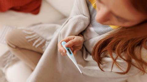 Eine kranke junge Frau, in eine Decke gehüllt, hält ein Fieberthermometer in der Hand, während sie auf einem weißen Sofa zu Hause sitzt.