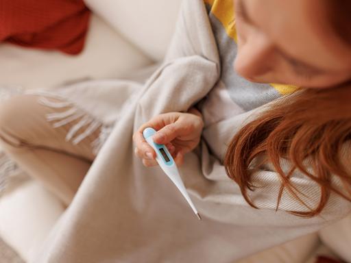 Eine kranke junge Frau, in eine Decke gehüllt, hält ein Fieberthermometer in der Hand, während sie auf einem weißen Sofa zu Hause sitzt.