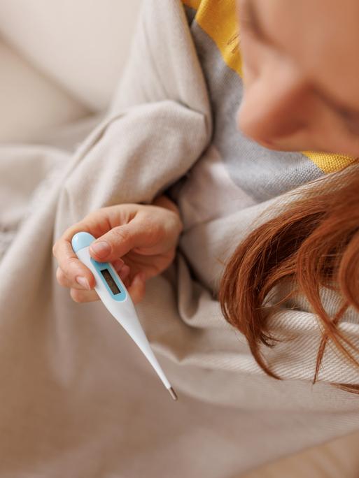 Eine kranke junge Frau, in eine Decke gehüllt, hält ein Fieberthermometer in der Hand, während sie auf einem weißen Sofa zu Hause sitzt.