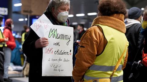 Eine Frau mit Atemschutzmaske trägt ein Schild vor dem Bauch, auf dem sie in englischer Sprache eine Unterkunft für eine Mutter mit kleinen Kindern anbietet. Mit dem Rücken zur Kamera steht vor ihr eine dunkelhäutige Frau, die eine gelbe Warnweste trägt.