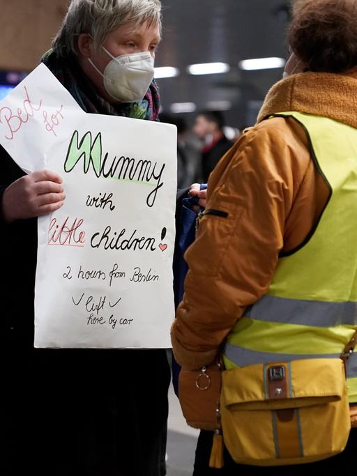 Eine Frau mit Atemschutzmaske trägt ein Schild vor dem Bauch, auf dem sie in englischer Sprache eine Unterkunft für eine Mutter mit kleinen Kindern anbietet. Mit dem Rücken zur Kamera steht vor ihr eine dunkelhäutige Frau, die eine gelbe Warnweste trägt.