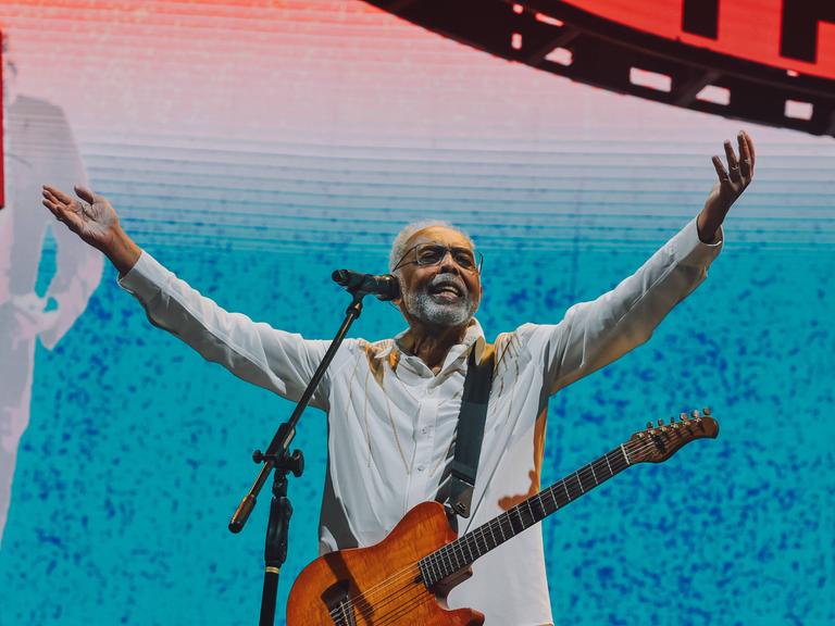 Der brasilianische Musiker Gilberto Gil steht mit einer Gitarre auf der Bühne. Er hält beide Arme in die Höhe und lacht.