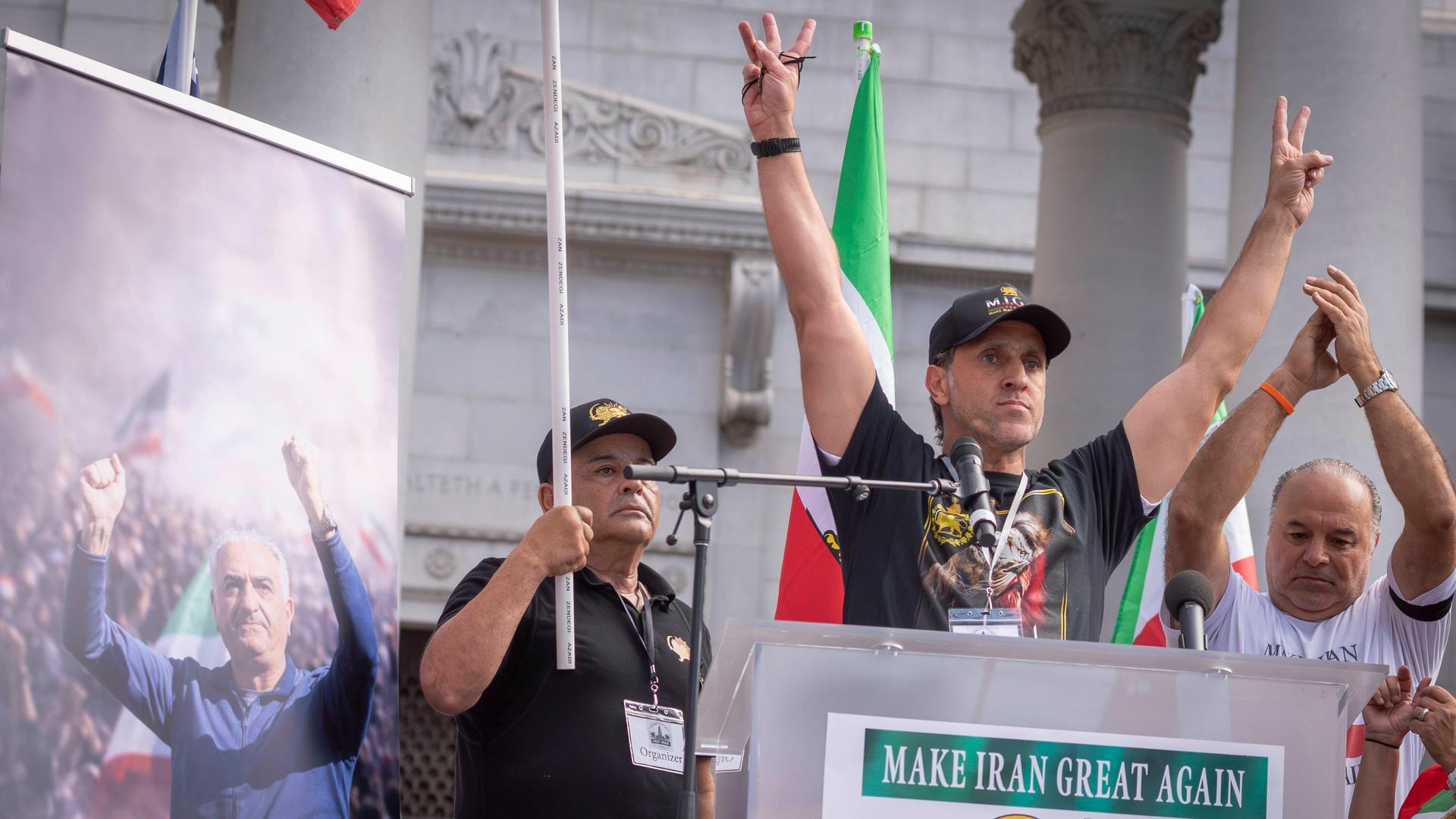 Der frühere Kapitän der iranischen Fußball-Nationalmannschaft Ali Karimi bei einer Demonstration von Exil-Iranern in Los Angeles. Er steht an einem Redepult mit der Aufschrift "Make Iran great again" und hebt beide Arme zum Victory-Zeichen nach oben.