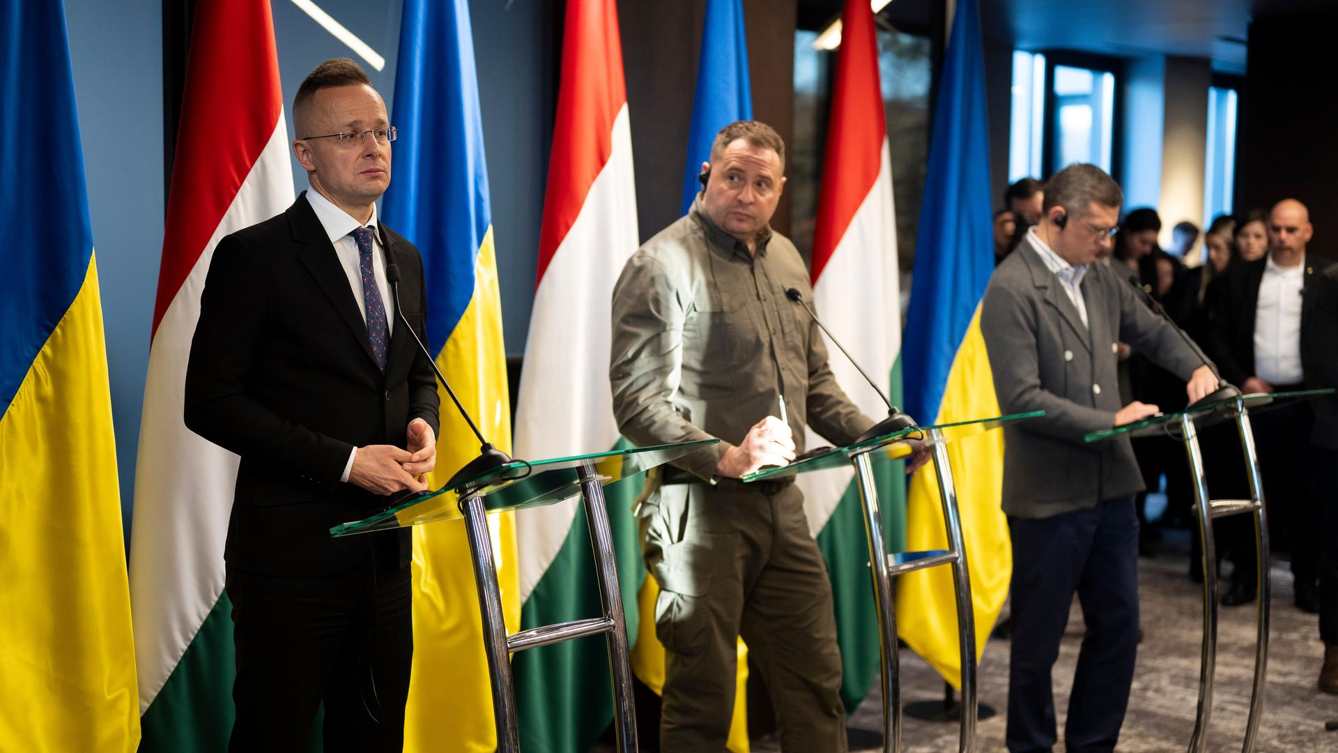 Die beiden Außenminister umrahmen eine dritte Person vor den Flaggen Ungarns und der Ukraine. Sie stehen an Rednerpulten. Die beiden Außenminister umrahmen eine dritte Person vor den Flaggen Ungarns und der Ukraine. Sie stehen an Rednerpulten.