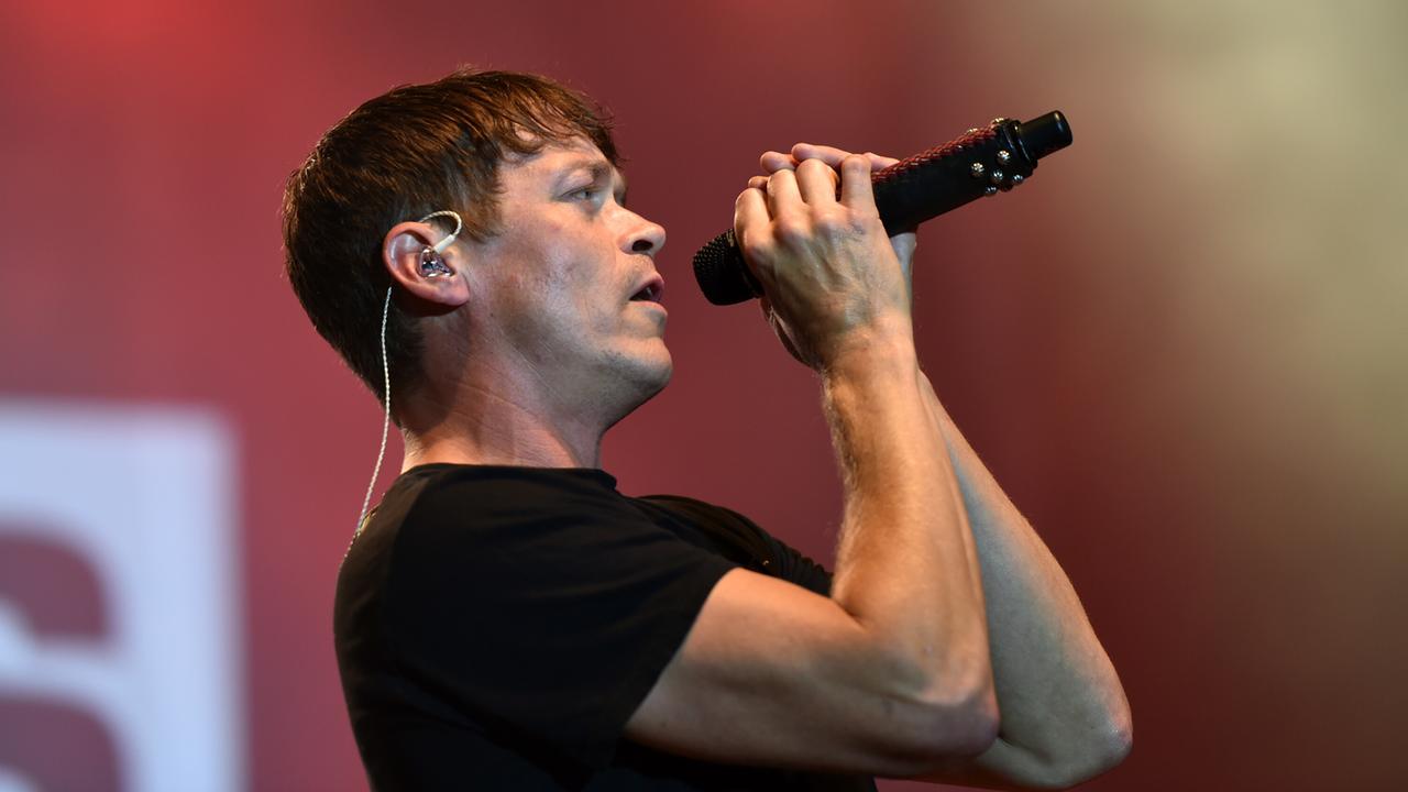 Man sieht Arnold von der Seite, wie er das Mikrofon mit beiden Händen an den Mund hält. Dahinter unscharf ein roter Bühnenhintergrund.