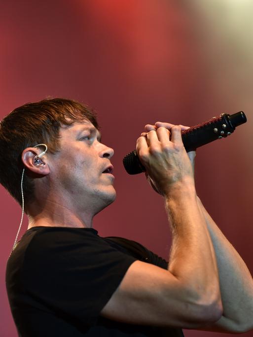 Man sieht Arnold von der Seite, wie er das Mikrofon mit beiden Händen an den Mund hält. Dahinter unscharf ein roter Bühnenhintergrund.