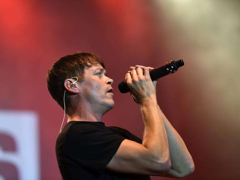 Man sieht Arnold von der Seite, wie er das Mikrofon mit beiden Händen an den Mund hält. Dahinter unscharf ein roter Bühnenhintergrund.