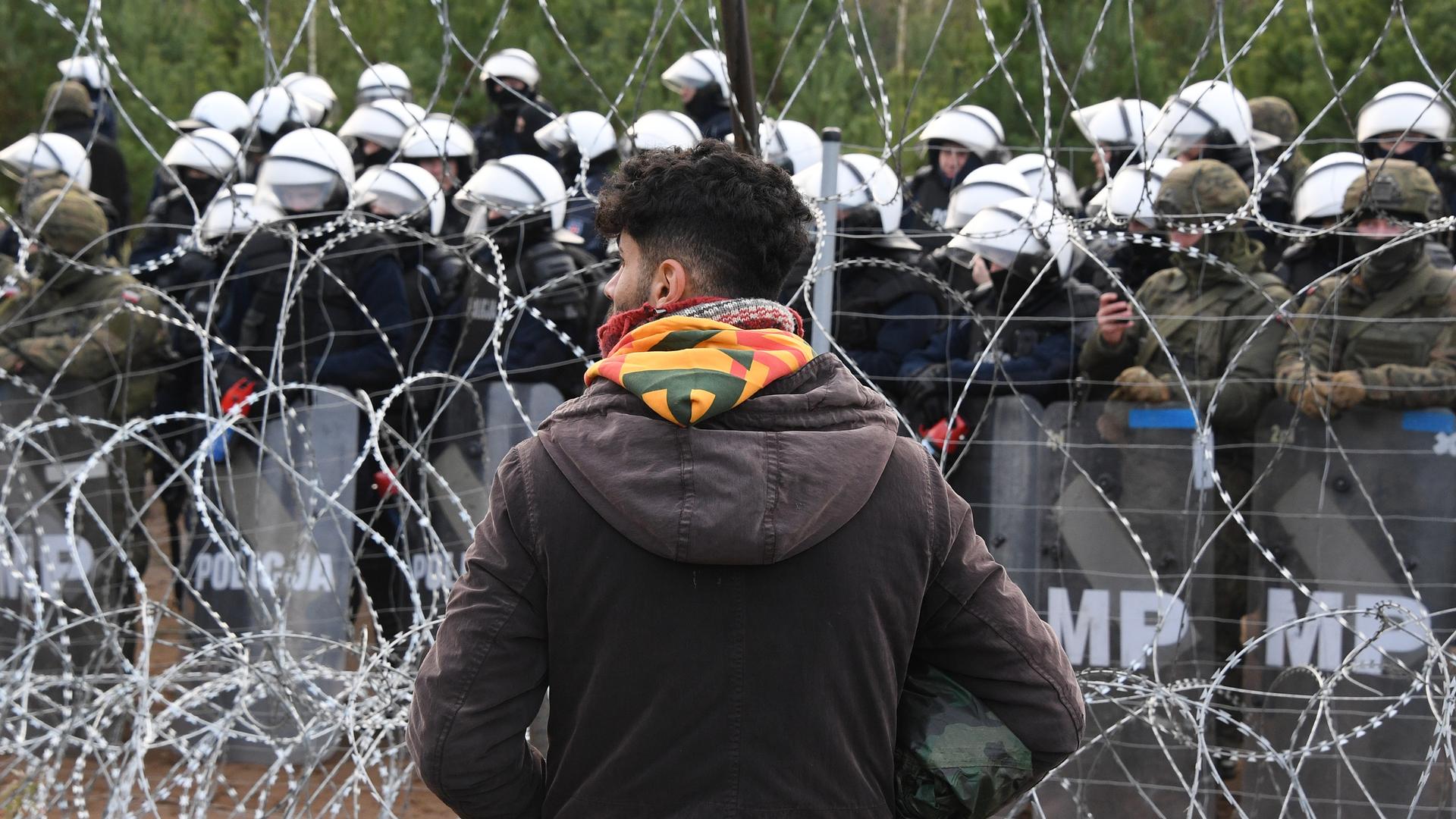 Ein Geflüchteter steht vor einem Stacheldrahtzaun und dutzenden Polizisten an belarusisch-polnischen Grenze. 