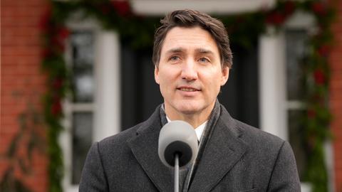 Justin Trudeau, ein Mann Mitte 50 mit dunklem Haar steht im Wintermantel vor einem Rednerpult vor einem Haus und spricht in ein Mikrofon. 