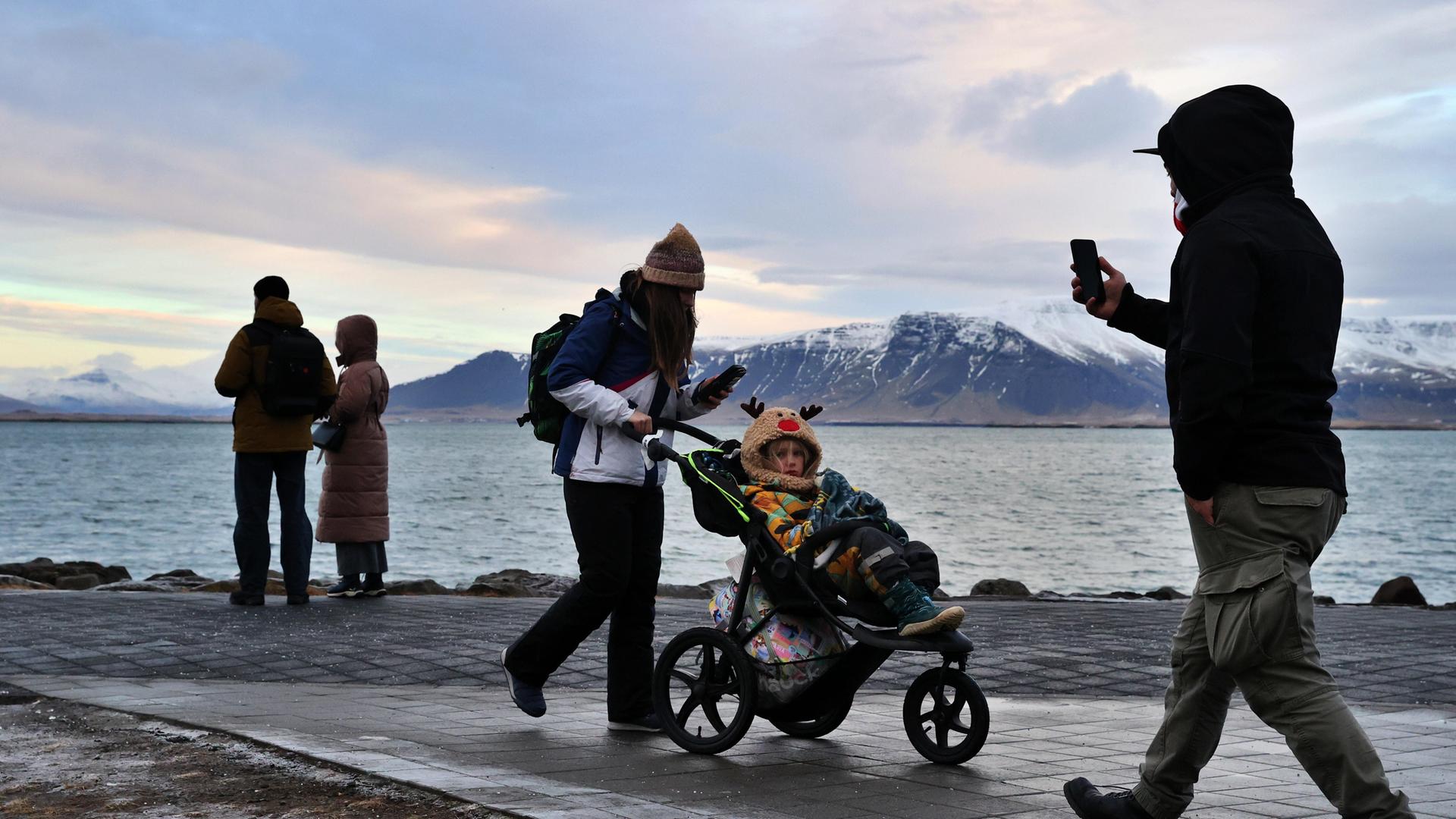 Menschen an einer Uferpromenade in Reykjavík, eine Frau schiebt einen Kinderwagen. Dahinter Meer und schneebedeckte Berge.
