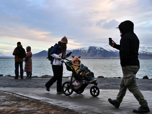 Menschen an einer Uferpromenade in Reykjavík, eine Frau schiebt einen Kinderwagen. Dahinter Meer und schneebedeckte Berge. Menschen an einer Uferpromenade in Reykjavík, eine Frau schiebt einen Kinderwagen. Dahinter Meer und schneebedeckte Berge.