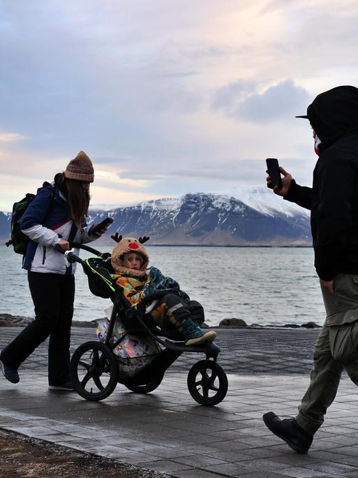 Menschen an einer Uferpromenade in Reykjavík, eine Frau schiebt einen Kinderwagen. Dahinter Meer und schneebedeckte Berge. Menschen an einer Uferpromenade in Reykjavík, eine Frau schiebt einen Kinderwagen. Dahinter Meer und schneebedeckte Berge.