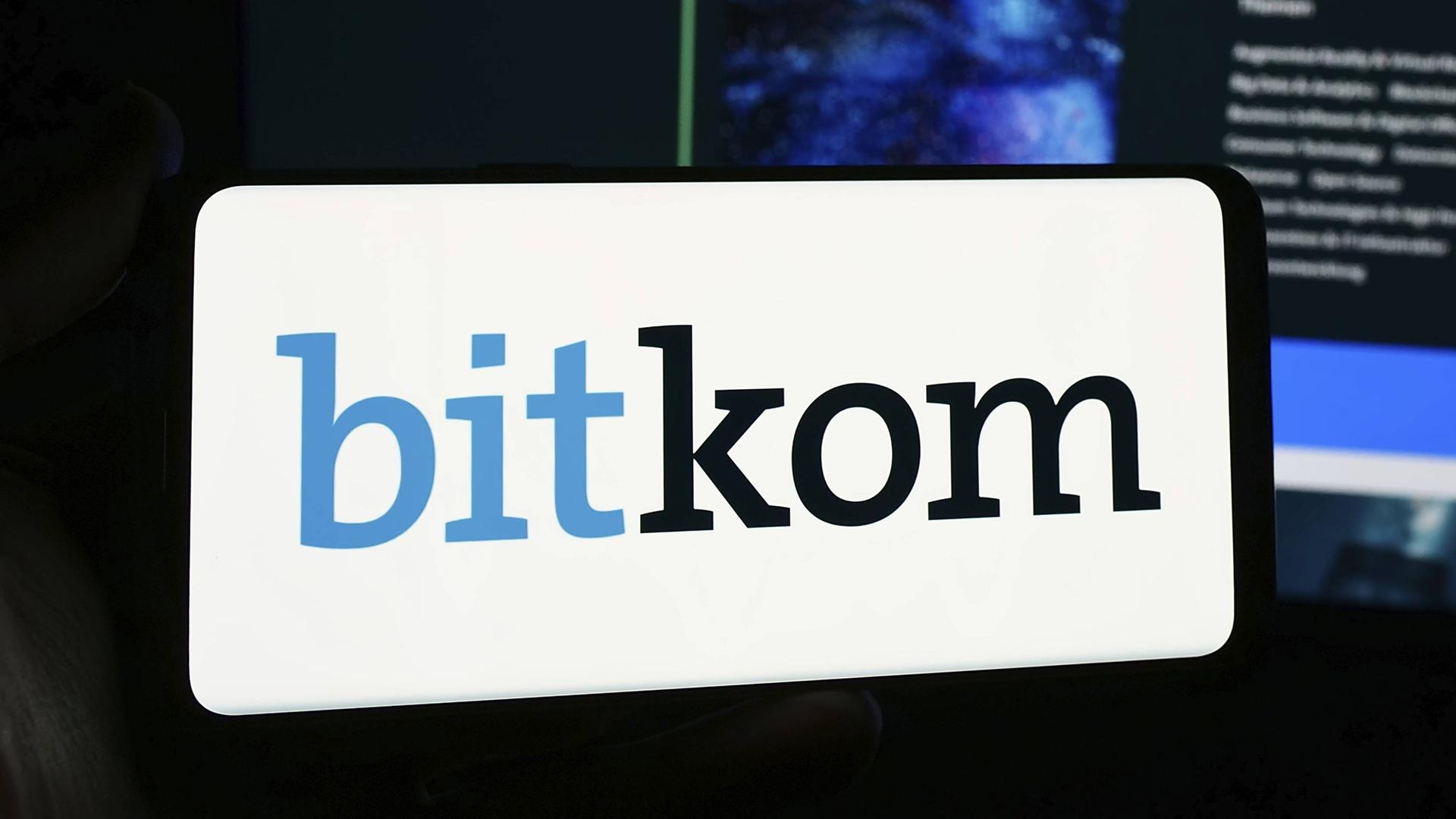 Eine Person hält ein Smartphone mit Logo des deutschen Telekommunikationsverbands Bitkom e. V. vor eine Website. 