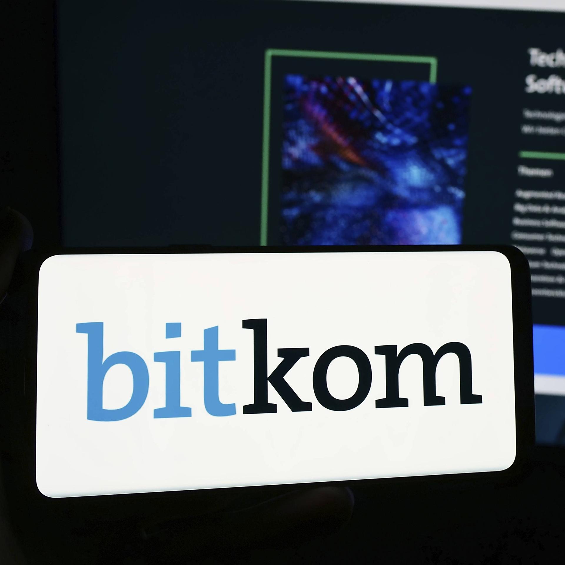 Eine Person hält ein Smartphone mit Logo des deutschen Telekommunikationsverbands Bitkom e. V. vor eine Website. 