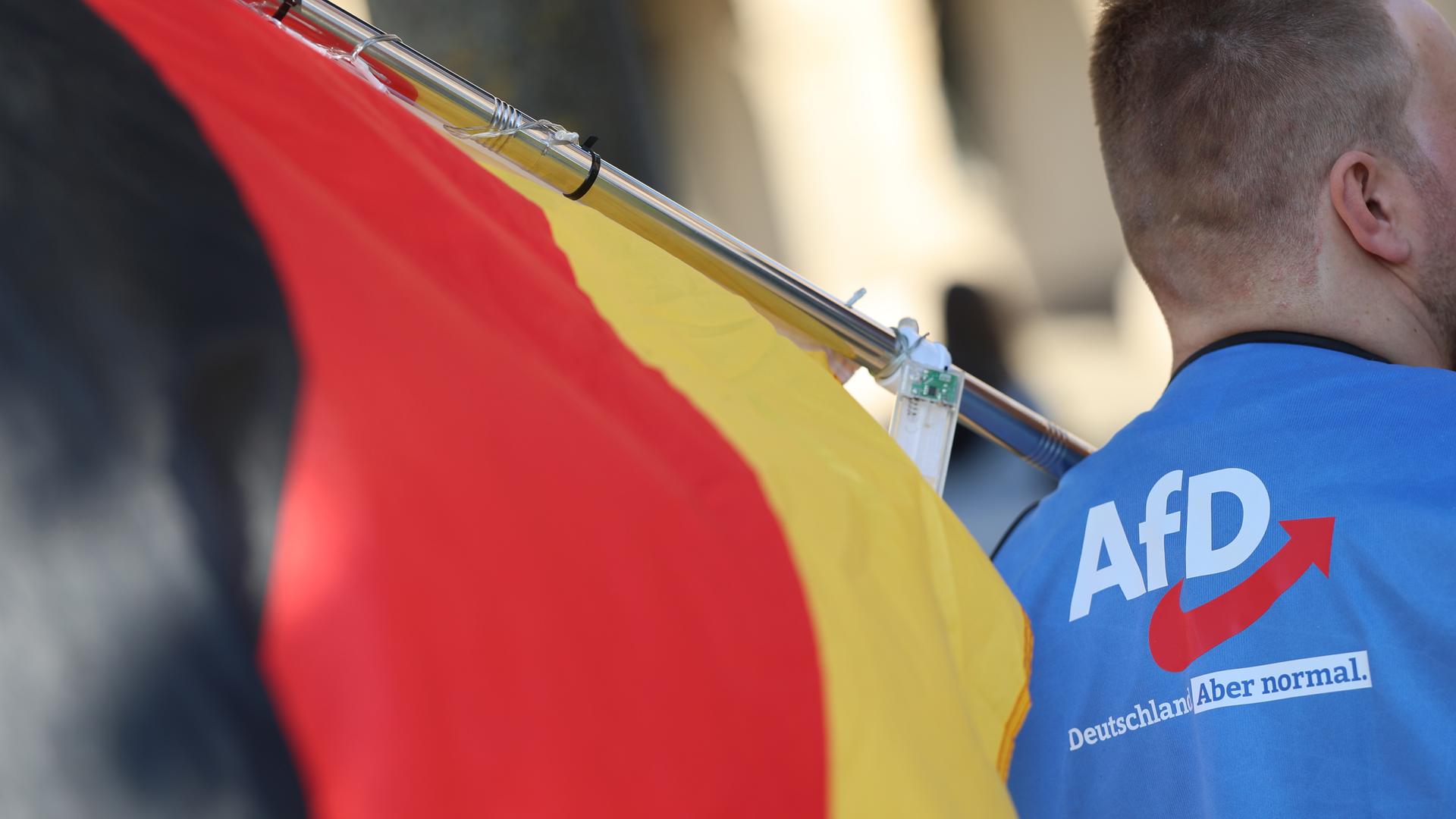Ein Mann trägt bei einer Demonstration ein T-Shirt mit einem AfD-Logo. Auf dem T-Shirt steht:  "Deutschland. Aber normal." Der Mann hält eine Deutschland-Fahne.