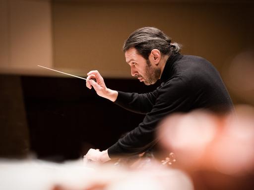 Vladimir Jurowski gibt gebeugt seinen Musikern mit dem Dirigierstab ein Zeichen für den Einsatz.