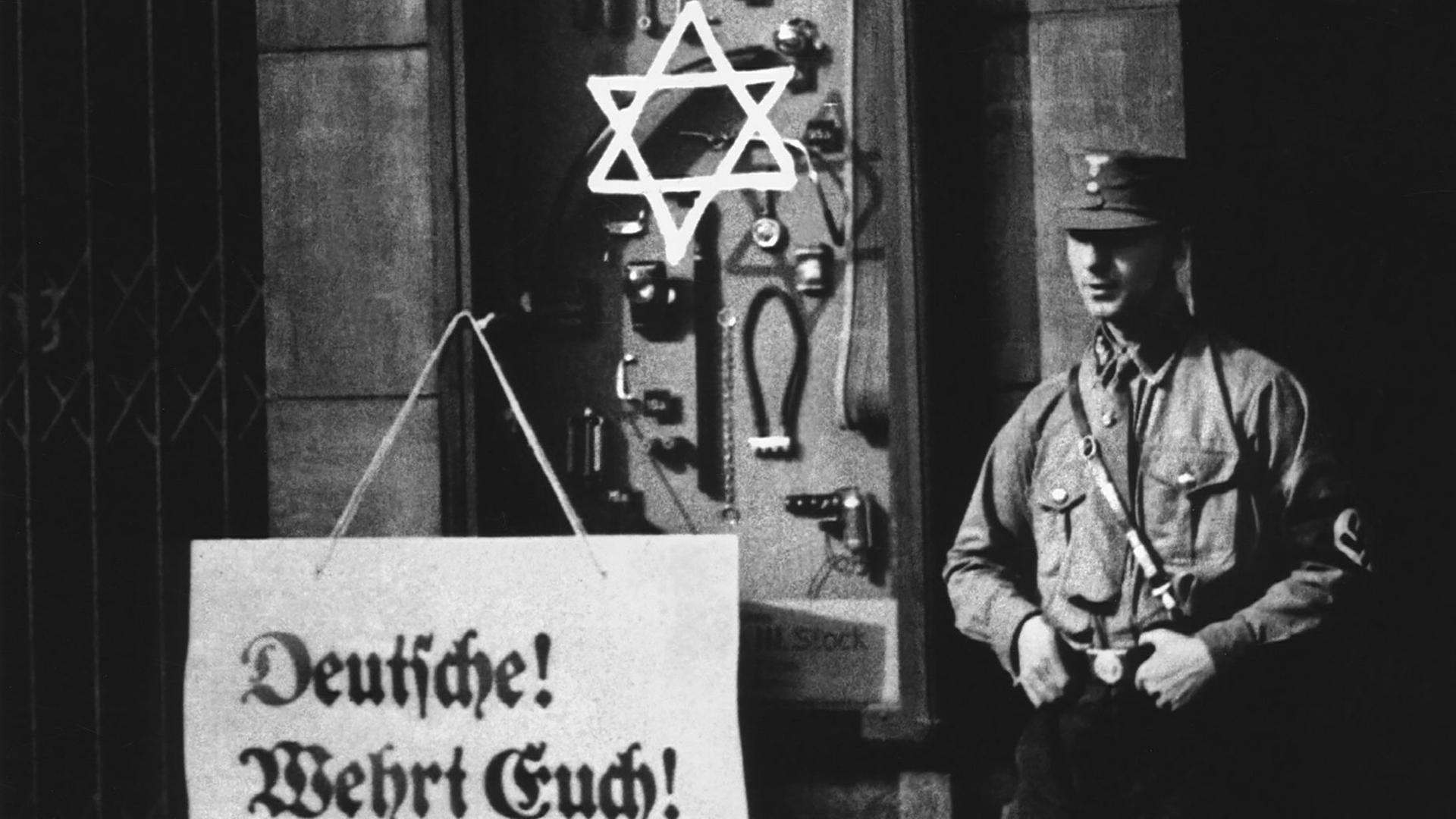 An einem Schaufenster steht auf einem Plakat: "Deutsche! Wehrt Euch! Kauft nicht bei Juden!", ein Aufruf zum Boykott jüdischer Geschäfte durch die Nationalsozialisten. Ein Judenstern ist auf das Schaufenster geschmiert, ein "Braunhemd" der SA (Sturmabteilung) steht als Wachposten daneben, Berlin, 1. April 1933