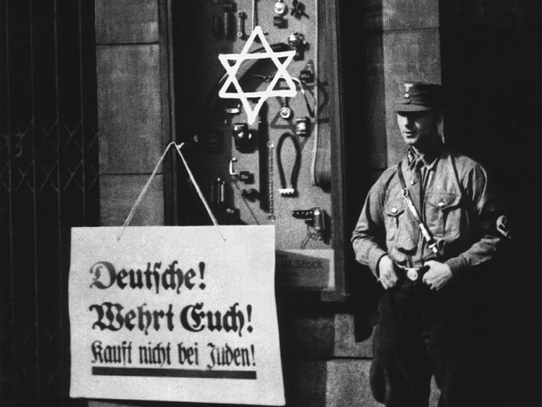 An einem Schaufenster steht auf einem Plakat: "Deutsche! Wehrt Euch! Kauft nicht bei Juden!", ein Aufruf zum Boykott jüdischer Geschäfte durch die Nationalsozialisten. Ein Judenstern ist auf das Schaufenster geschmiert, ein "Braunhemd" der SA (Sturmabteilung) steht als Wachposten daneben, Berlin, 1. April 1933