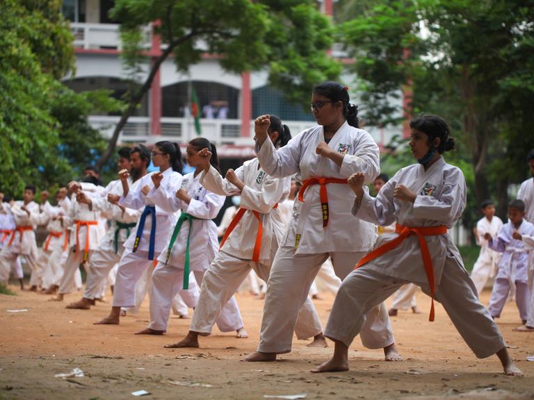 Das Bild zeigt Mädchen aus Bangladesch, die draußen in weißen Karateanzügen und unterschiedlichen Gürtelfarben Karate trainieren.