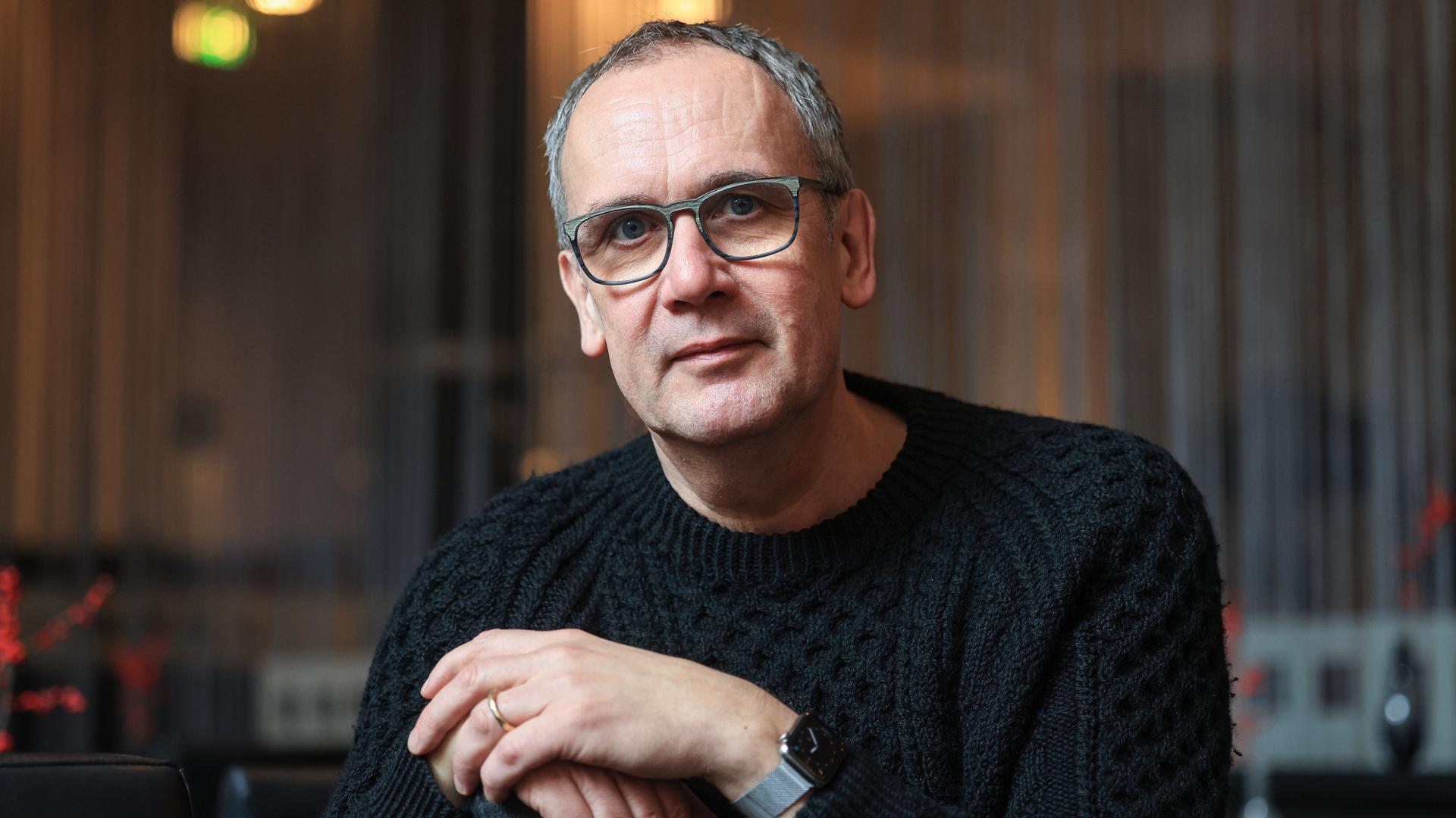 Volker Kutscher schaut in die Kamera. Er trägt eine große Brille und einen schwarzen Grobstrickpullover.