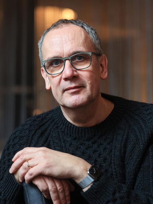 Volker Kutscher schaut in die Kamera. Er trägt eine große Brille und einen schwarzen Grobstrickpullover.