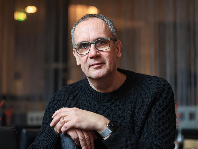 Volker Kutscher schaut in die Kamera. Er trägt eine große Brille und einen schwarzen Grobstrickpullover.