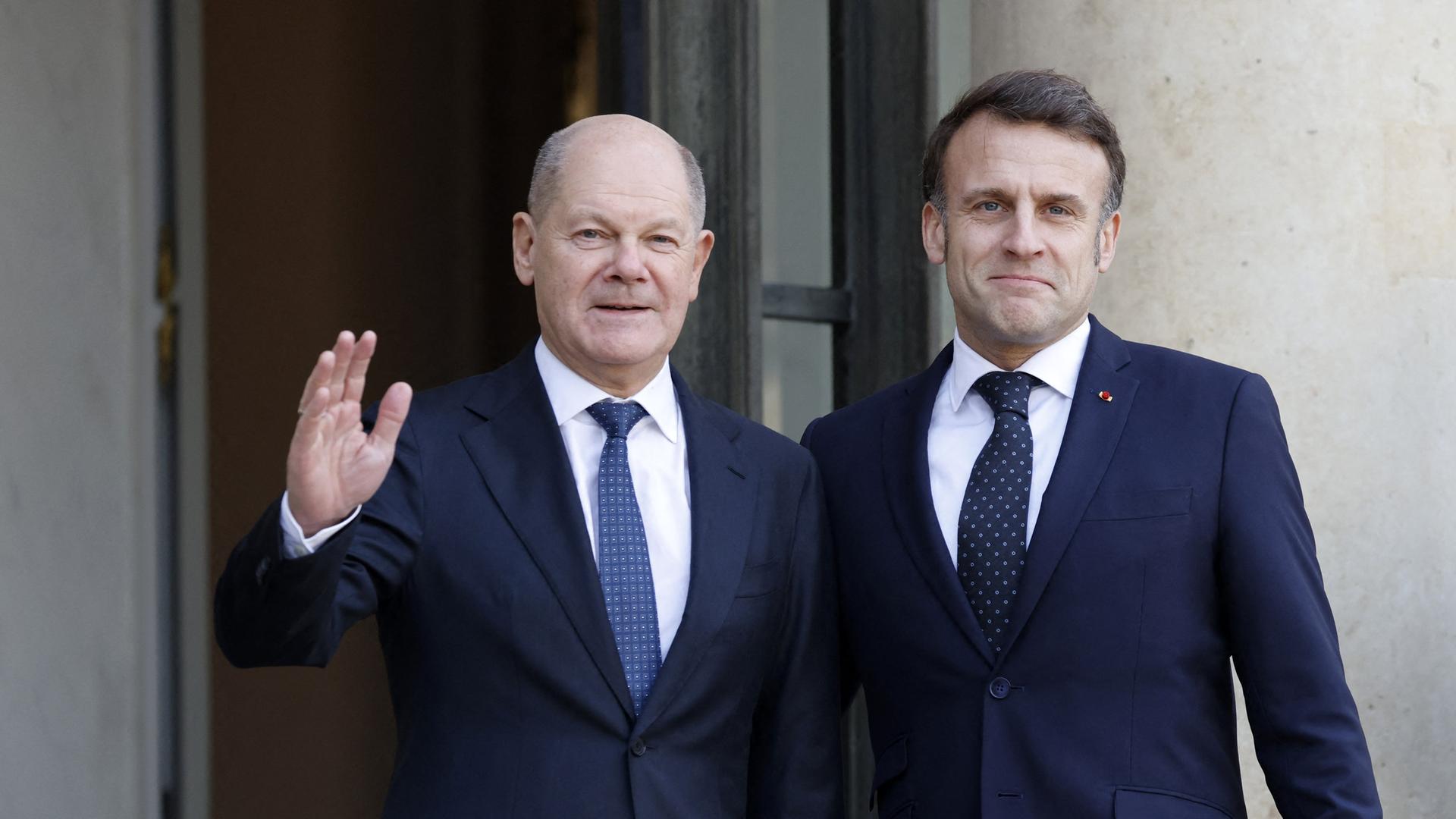 Bundeskanzler Scholz steht vor dem Elysee-Palast neben dem französischen Staatspräsidenten Marcon. Beide tragen dunkelblaue Anzüge, Scholz hebt freundlich grüßend die rechte Hand. 