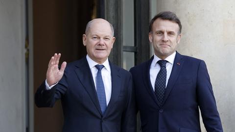 Bundeskanzler Scholz steht vor dem Elysee-Palast neben dem französischen Staatspräsidenten Marcon. Beide tragen dunkelblaue Anzüge, Scholz hebt freundlich grüßend die rechte Hand. Aufnahme vom 17.02.2025.