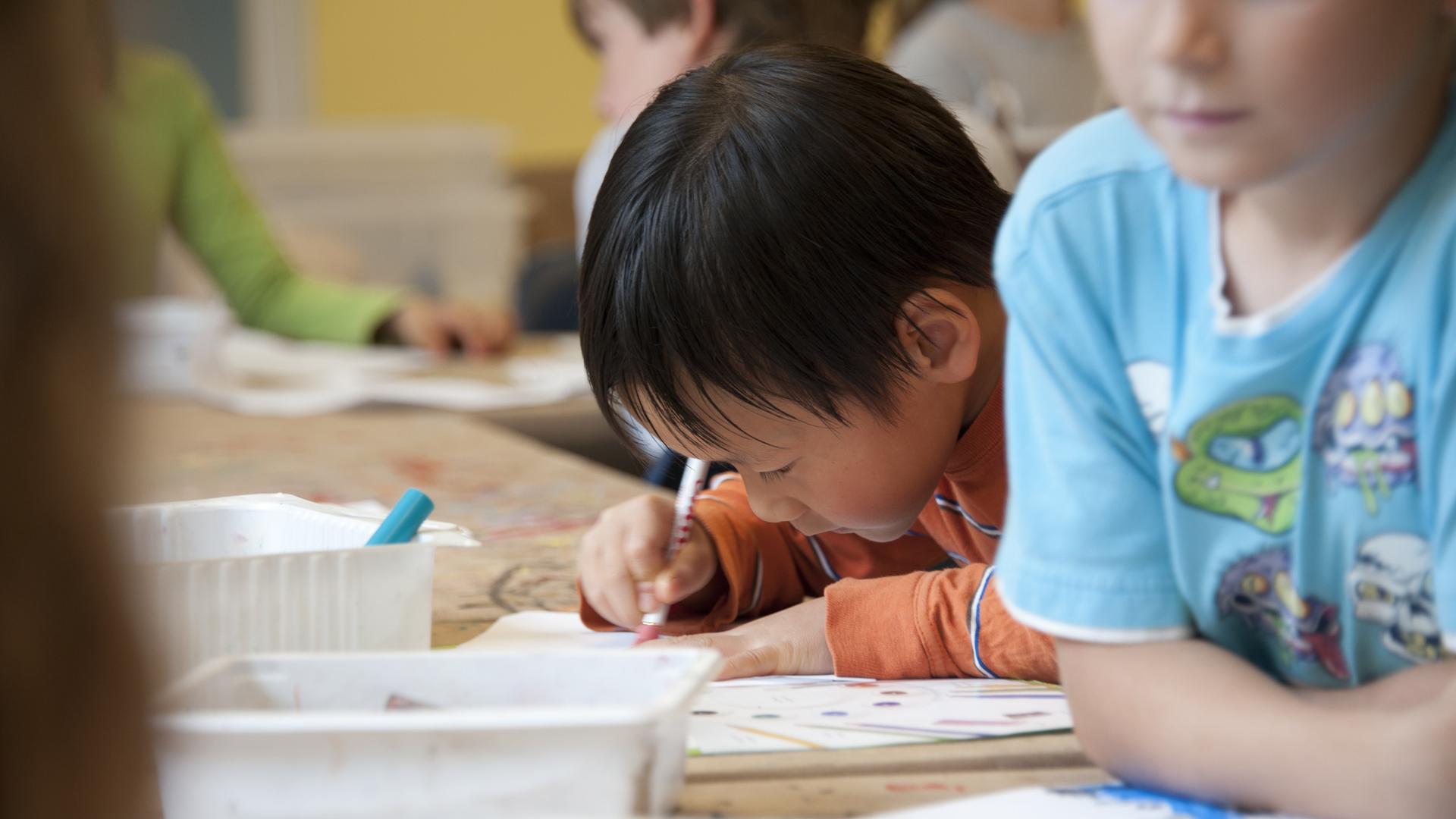 Kinder in einem Klassenzimmer. Im Bildzentrum ist ein Kind mit dunklen Haaren und orangefarbenem Shirt. Es malt.