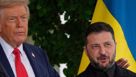 Nahaufnahme der Oberkörper der beiden nebeneinander vor einer ukrainischen Flagge. Trump legt seine Hand auf Selenskyjs Schulter.