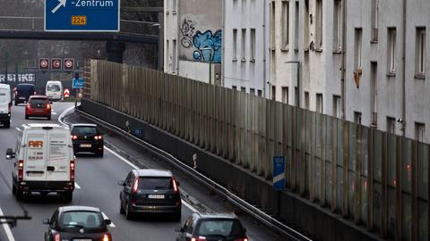 Im Vordergrund sihet man eine stark befahrene Autobahn. Dierekt neben der Autobahn, abgetrennt durch Lärmschutzwende, befinden sich Wohnhäuser.