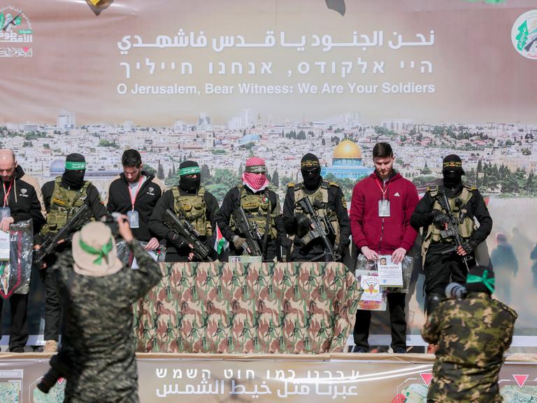 Drei Geiseln stehen, umgeben von Hamas-Terroristen, auf einer Bühne. Es werden Fotos gemacht. Die Terroristen tragen Uniformen und sind vermummt. Die Geiseln sind in zivil und unvermummt.