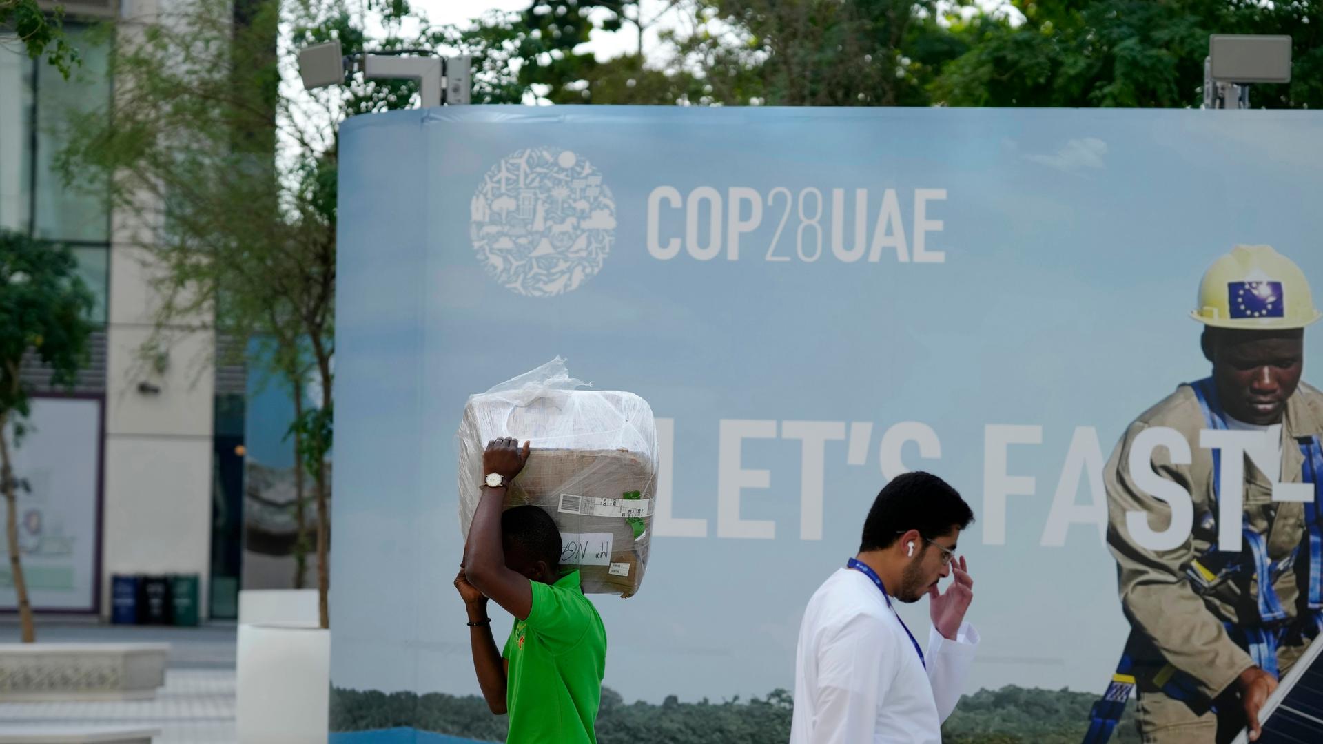 Menschen laufen in Dubai vor einem Plakat des UN-Klimagipfels COP28 in entgegengesetzte Richtungen.